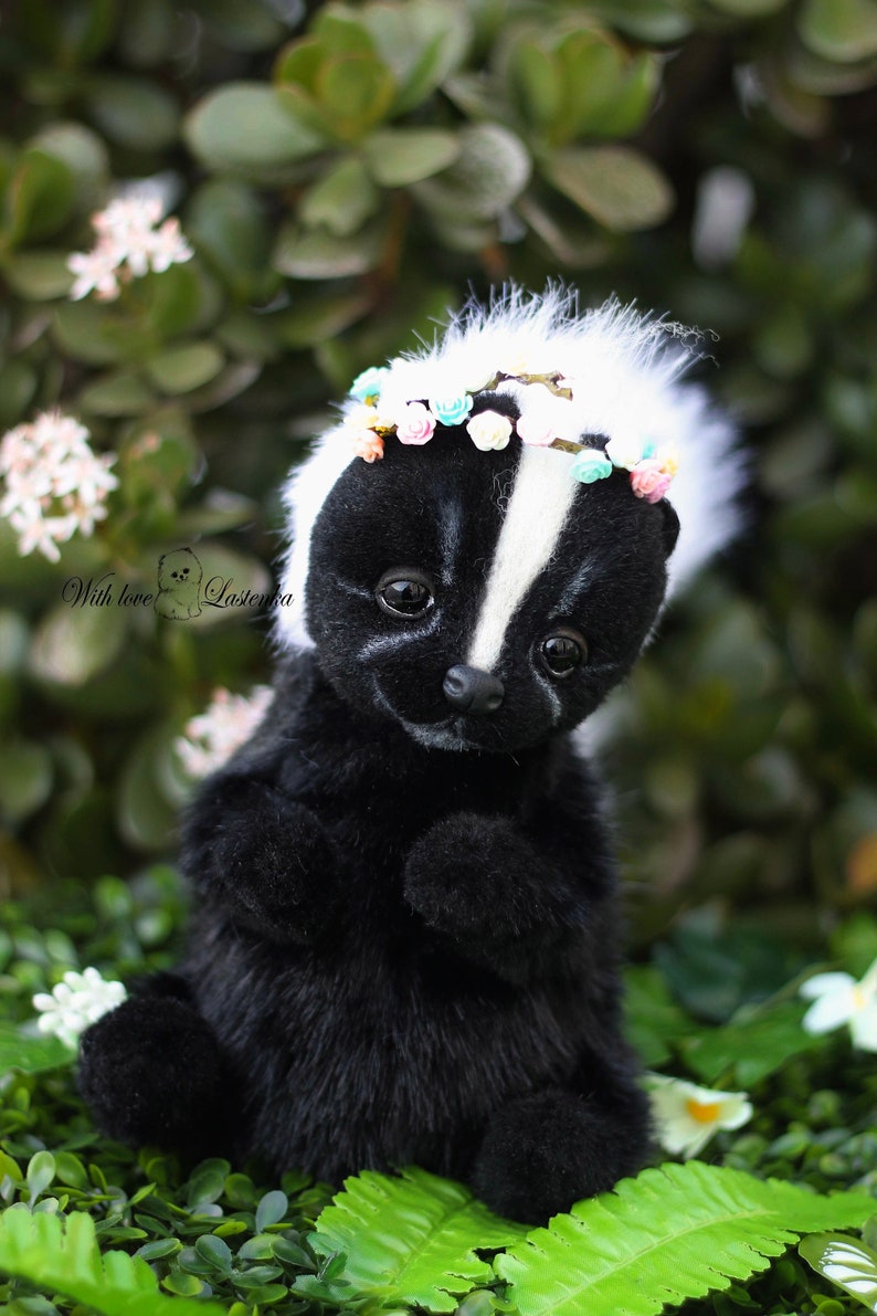 May include: A black and white skunk plush toy wearing a flower crown. The skunk is sitting on a bed of green leaves.