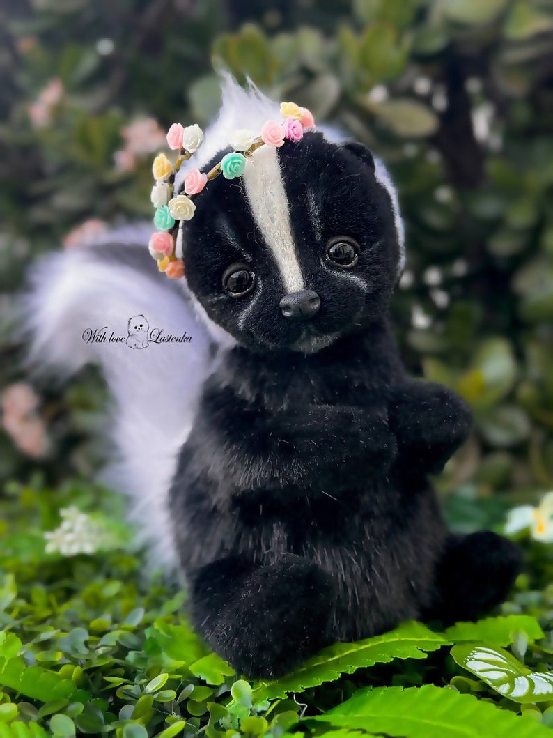 May include: A black and white skunk plush toy wearing a colorful flower crown. The skunk is sitting on a bed of green leaves. "With love, Losenka" is written on the plush toy.