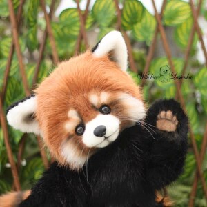 Puede incluir: Un peluche de panda rojo con una cabeza marr&oacute;n rojiza y esponjosa, orejas blancas y un cuerpo negro. El panda rojo est&aacute; saludando con su pata. El fondo presenta un enrejado verde y marr&oacute;n.