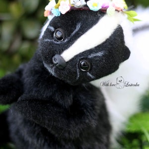 May include: A black and white skunk plush toy wearing a flower crown. The skunk is looking up with its eyes open and has a white stripe down its back. The toy is sitting in green grass.