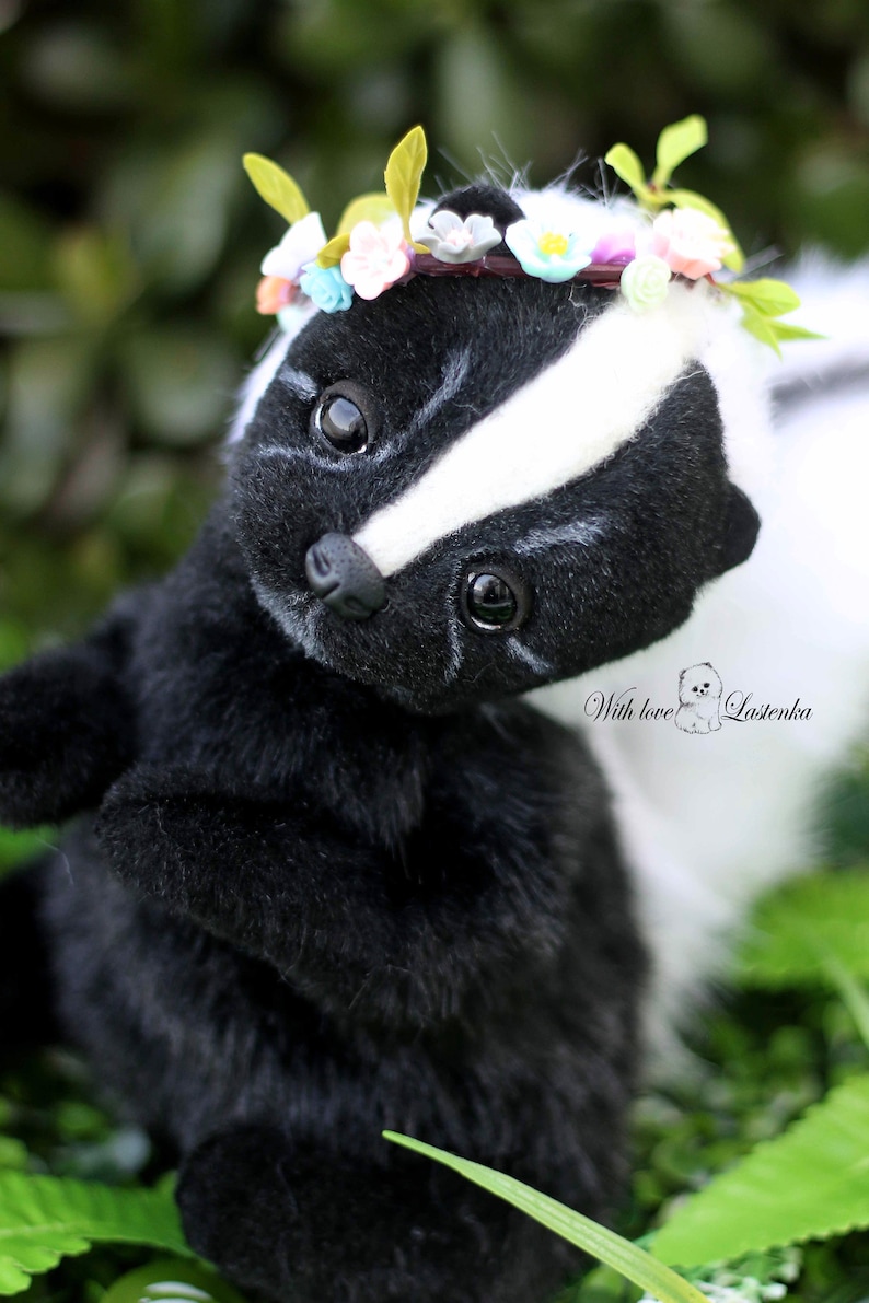 May include: A black and white skunk plush toy wearing a flower crown. The skunk is looking up with its eyes open and has a white stripe down its back. The toy is sitting in green grass.