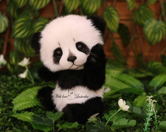 May include: A plush panda bear with black and white fur, sitting amidst green foliage and white flowers. The panda has a white face, black eyes, and black ears and limbs. The text "With love...Lazenka" is on the panda's chest.