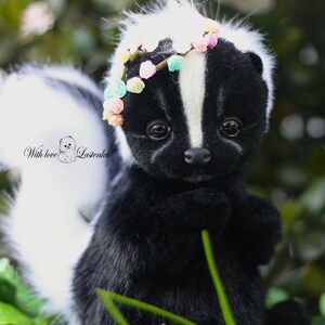 May include: A black and white skunk plush toy wearing a flower crown. The skunk is sitting in a bed of green grass with white flowers.
