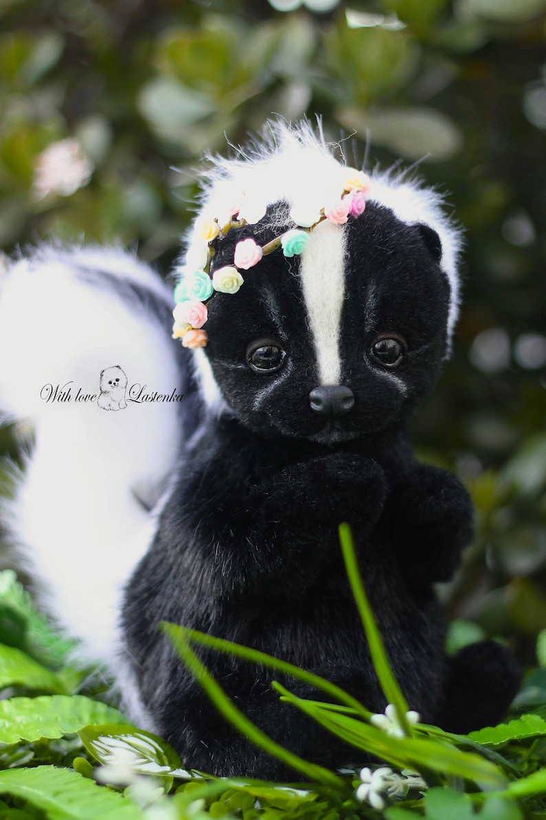 May include: A black and white skunk plush toy wearing a flower crown. The skunk is sitting in a bed of green grass with white flowers.