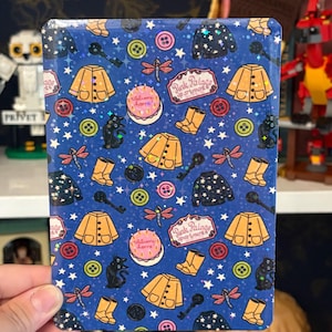 May include: A rectangular sticker with a dark blue background featuring a repeating pattern of yellow raincoats, yellow rain boots, pink cakes with the text Welcome home, black cats, pink dragonflies, buttons, keys, and sweaters with stars.