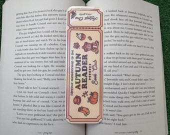 May include: A rectangular paper bookmark with a cream background and a brown border. The text reads Admit One, Autumn Reader Book Club, and lifetime membership to the. Illustrations include a bear, a cup, a pumpkin, a sock, and leaves.