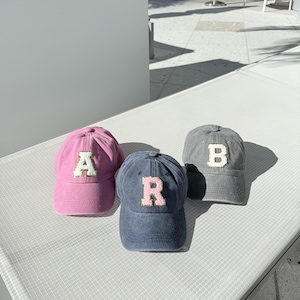 May include: Three baseball caps in pink, navy blue, and gray. Each cap features a different letter: "A", "R", and "B" respectively, in a contrasting color. The caps are displayed on a white surface, with outdoor seating in the background.
