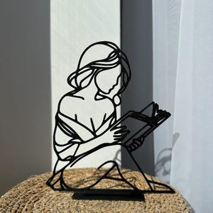 Può includere: Silhouette nera in metallo di una persona che legge un libro. La figura è seduta, con il libro aperto tra le mani. L'opera d'arte è posta su una superficie tessuta, con una tenda bianca sullo sfondo. La silhouette è un oggetto decorativo.