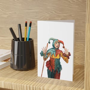 Puede incluir: Una tarjeta de felicitación en blanco con una ilustración en acuarela de un bufón con un disfraz colorido. El bufón sostiene un bastón de madera y tiene las manos levantadas. La tarjeta está sobre una superficie de madera con un portalápices negro y lápices.