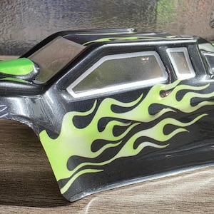 Può includere: Una carrozzeria per auto radiocomandata nera con grafiche a fiamma verde lime brillante sui lati e sul cofano, uno spoiler posteriore verde lime abbinato e sezioni trasparenti per i finestrini. L'esterno ha una finitura lucida.