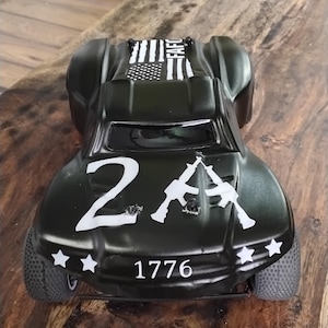 Può includere: Macchina giocattolo nera con grafica bianca, tra cui il numero "2A", due fucili incrociati, l'anno "1776" e un disegno della bandiera americana. L'auto ha un design aerodinamico e pneumatici visibili.