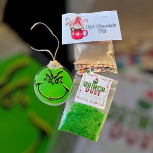 May include: A festive holiday-themed gift set featuring a green Grinch-faced ornament, a packet of green Grinch Dust, and a hot chocolate kit. The hot chocolate kit includes a packet of mix, sprinkles, and small marshmallows. The ornament is round and transparent.