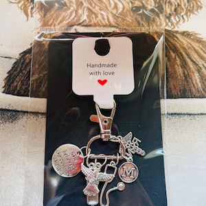 Peut inclure: Un porte-clés argenté avec plusieurs breloques, dont un ange, un stéthoscope et un disque avec l'inscription "She believed she could so she did". Le porte-clés comporte également un charm avec la lettre "M" et les mots "RN Nurse". L'étiquette indique "Handmade with love".