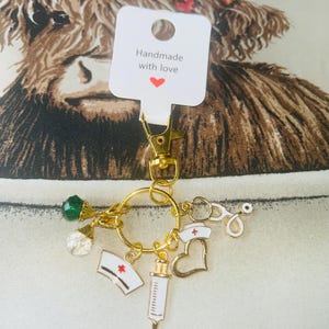 May include: Gold-coloured keychain with medical charms: a nurse's cap, syringe, and stethoscope. The keychain also has a green and clear crystal charm. A tag reads "Handmade with love."
