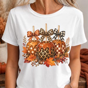 May include: A white short-sleeve t-shirt featuring a graphic print of three caramel apples. The apples are decorated with polka dot, leopard, and checkered patterns, each topped with a matching bow and surrounded by autumn leaves and berries.