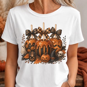 Pode incluir: Uma t-shirt branca com uma estampa gráfica de três maçãs do amor com laços, um pequeno fantasma, uma abóbora, um chapéu de bruxa e um crânio. O design inclui folhas de outono e bagas, criando um tema sazonal festivo na parte da frente.