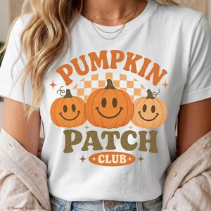 Pode incluir: Uma t-shirt branca com um gráfico de três abóboras cor de laranja com rostos sorridentes. O texto diz PUMPKIN PATCH CLUB em letras cor de laranja e verde-azeitona. O design inclui um padrão de fundo axadrezado e pequenas estrelas.