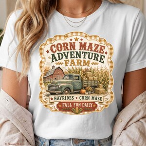 Pode incluir: Uma t-shirt branca com um gráfico de uma carrinha verde antiga carregada com fardos de feno em frente a um celeiro vermelho e um milharal. O texto diz CORN MAZE ADVENTURE FARM, HAYRIDES, CORN MAZE e FALL FUN DAILY numa moldura decorativa.