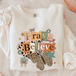 Puede incluir: Sudadera color crema con las palabras "Pray & Believe" en un diseño colorido de letras en bloque. El diseño incluye una mariposa con estampado de leopardo, estrellas y lazos. La sudadera es de un material suave.