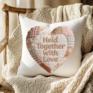 May include: A white square pillow with a heart-shaped patch. The patch is made of various fabrics in shades of pink, brown, and cream, with floral and gingham patterns. The text "Held Together With Love" is embroidered on the heart.