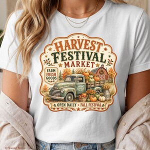 Pode incluir: Uma t-shirt branca com uma estampa gráfica contendo o texto Harvest Festival Market, Farm Fresh Goods, Open Daily e Fall Festival. O design inclui uma carrinha pick-up verde vintage carregada de abóboras e um celeiro vermelho ao fundo.