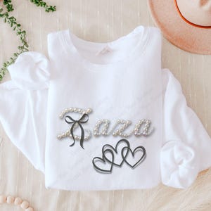 May include: A white long-sleeved sweatshirt with the name "Saza" in pearl-like letters, a black bow, and three interlocking hearts. The design is centered on the front of the shirt.