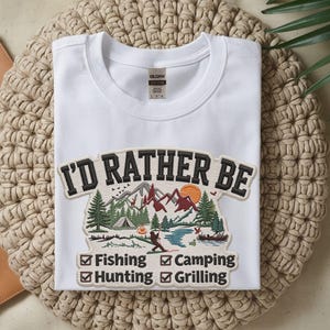 May include: White crew neck t-shirt with a graphic that reads "I'D RATHER BE" above a nature scene with mountains, trees, and a lake. Below the graphic are check boxes for fishing, camping, hunting, and grilling.
