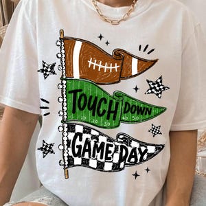 May include: A white t-shirt featuring a graphic of three stacked pennant flags. The top flag is brown with a football design, the middle green flag reads "TOUCH DOWN" with yard lines, and the bottom flag is black and white checkered with "GAME DAY".