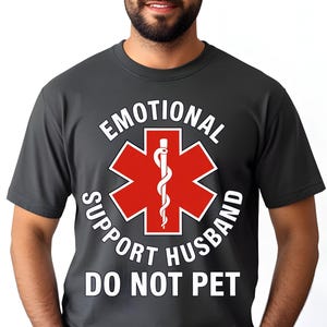 May include: Dark gray t-shirt with the text "EMOTIONAL SUPPORT HUSBAND DO NOT PET" in white. The shirt features a red star of life symbol with a white medical staff in the center.