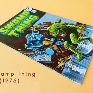 Op de afbeelding: Een vintage DC Comics Swamp Thing stripboek nummer 6 uit 1976. De cover toont een groen mensachtig wezen en een blauwe robotfiguur in een donkere kamer. De titel Swamp Thing staat in het geel gedrukt in de linkerbovenhoek.
