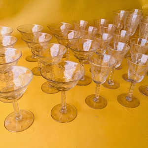 May include: A collection of clear glass stemware featuring etched leaf patterns on the bowls and twisted stems. These glasses include various shapes such as wide coupe glasses and taller, narrower goblets, all arranged together on a yellow surface.