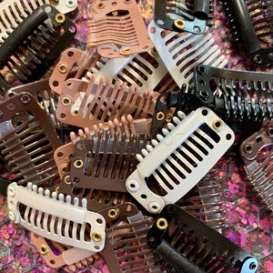 May include: A collection of hair extension clips in various colors, including black, white, and shades of brown. The clips have a comb-like design with small teeth and metal hardware for attachment. The background is a glittery, iridescent surface.