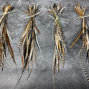 May include: Four bundles of decorative feathers in shades of brown, black, white, and tan, tied with twine. The feathers vary in length and pattern, creating a natural, textured look. The bundles are arranged on a gray surface.
