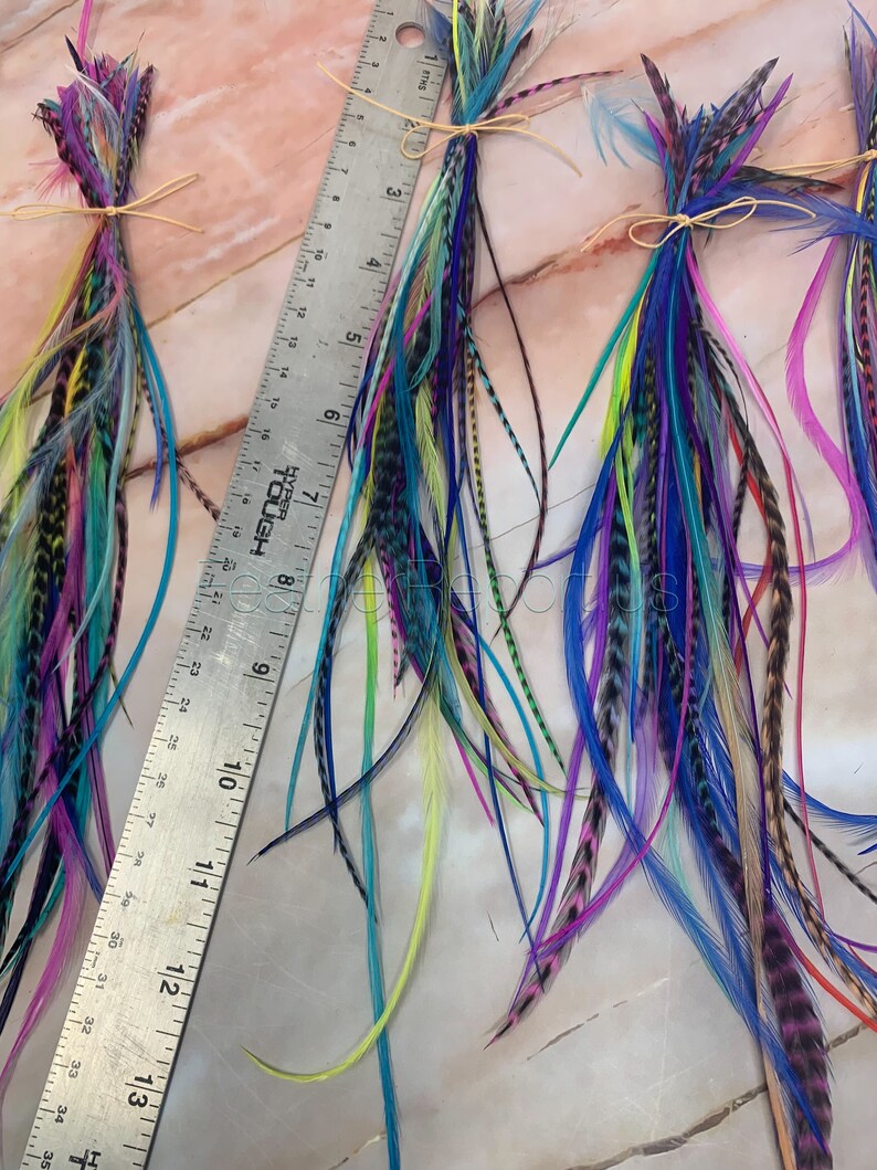 Puede incluir: Tres paquetes de plumas coloridas atadas con cuerda. Las plumas son de diferentes longitudes y colores, incluyendo azul, verde, morado, rosa y amarillo. Una regla est&aacute; en el fondo para la escala.