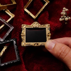 May include: A small, rectangular gold-colored picture frame with ornate, raised scrollwork detailing along the edges. The frame features a solid black center insert and is displayed alongside other similar miniature frames in gold, black, and clear.