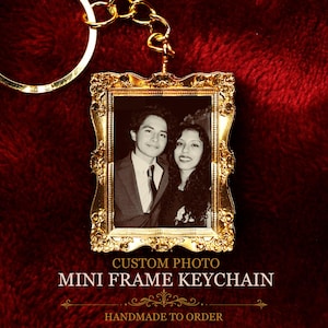 Op de afbeelding: Een goudkleurige metalen sleutelhanger met een rechthoekig frame met sierlijke, decoratieve randen. Het frame bevat een zwart-witfoto van twee personen. De tekst CUSTOM PHOTO MINI FRAME KEYCHAIN staat onder het frame.