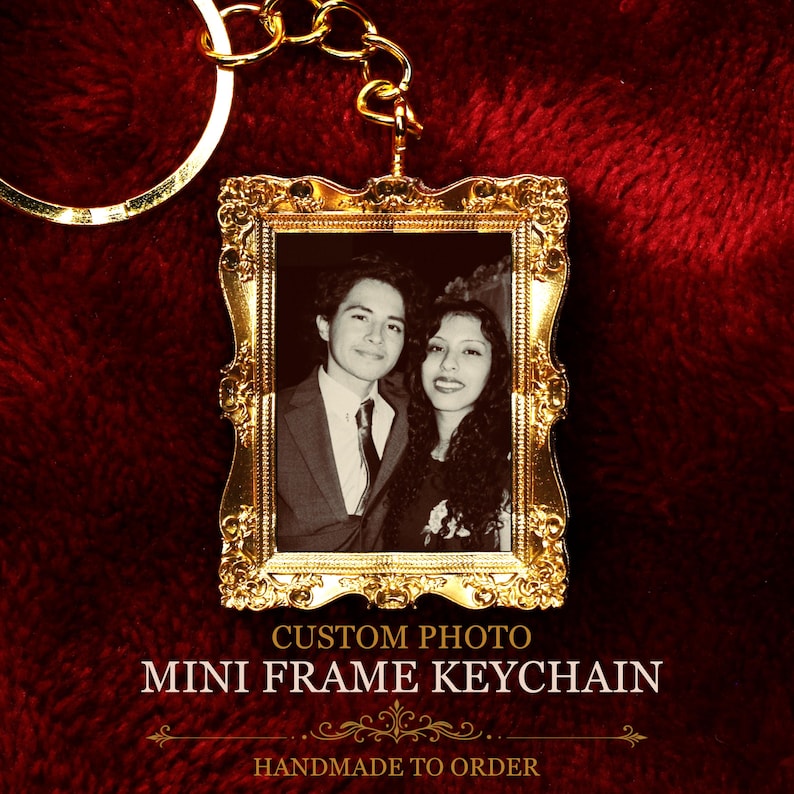 May include: A gold-toned metal keychain featuring a rectangular frame with ornate, decorative edges. The frame holds a black and white photograph of two people. The text CUSTOM PHOTO MINI FRAME KEYCHAIN is printed below the frame.