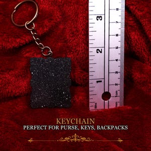 May include: A black rectangular keychain with a glittery finish attached to a bronze-colored metal ring and chain. A metal ruler placed next to the keychain shows it measures approximately 2.5 inches in height and 1.75 inches in width.