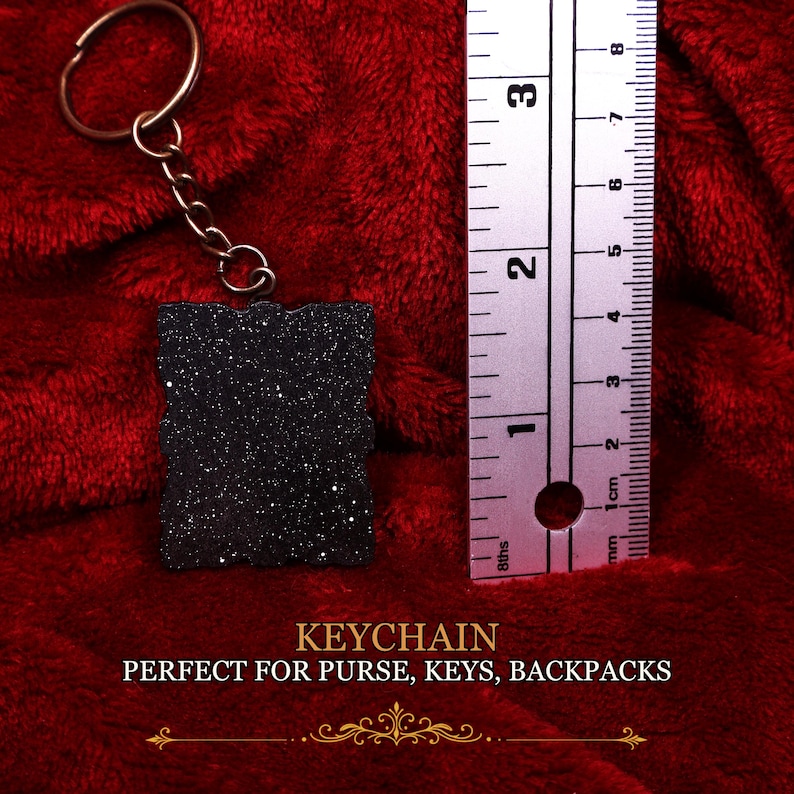 May include: A black rectangular keychain with a glittery finish attached to a bronze-colored metal ring and chain. A metal ruler placed next to the keychain shows it measures approximately 2.5 inches in height and 1.75 inches in width.