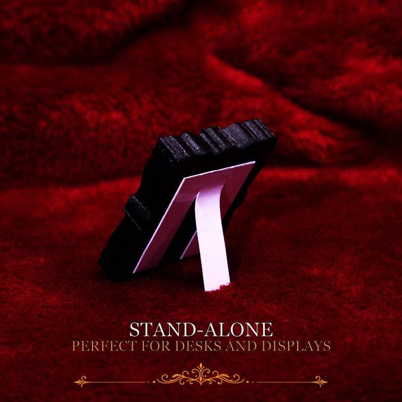 May include: A black rectangular object with a textured surface, supported by a white easel-style stand attached to the back. The text STAND-ALONE PERFECT FOR DESKS AND DISPLAYS is printed in white below the object against a deep red background.