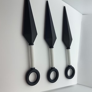 Peut inclure: Trois couteaux kunai noirs avec des manches blancs et des anneaux noirs à la base. Les lames sont tranchantes et angulaires, et les manches sont cylindriques. Les couteaux sont exposés sur un fond blanc.