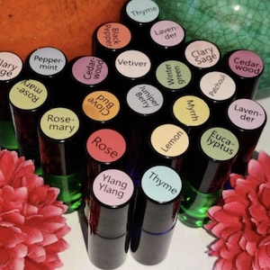 May include: A collection of dark glass roller bottles with colored labels featuring text including Thyme, Lavender, Clary Sage, Peppermint, Black Pepper, Cedarwood, Vetiver, Wintergreen, Rosemary, Clove Bud, Juniper Berry, Myrrh, Patchouli, Rose, Lemon, Eucalyptus, and Ylang Ylang.