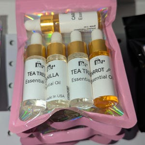 Könnte beinhalten: Eine rosa Tasche mit vier kleinen Glasflaschen mit ätherischen Ölen. Die Flaschen haben goldene Verschlüsse und weiße Etiketten mit schwarzem Text. Die Etiketten lauten "Tea Tree Essential Oil", "Vanilla Essential Oil" und "Carrot Essential Oil".