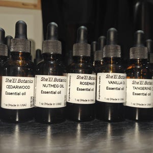 Puede incluir: Una colección de botellas de aceite esencial de vidrio ámbar con cuentagotas negros. Cada botella tiene una etiqueta blanca con el nombre del producto y la marca, incluyendo Cedro, Nuez moscada, Romero y Vainilla. Las botellas son de 30 ml.