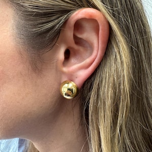 May include: A close-up of a gold-colored, round stud earring. The earring is a simple, polished sphere, reflecting the surrounding environment. The earring is worn on an ear with blonde and brown hair.