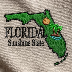 Op de afbeelding: Geborduurde patch met de groene omtrek van Florida, met een bruine vrucht en palmbomen. De tekst "FLORIDA Sunshine State" is zwart geborduurd. De patch bevindt zich op een beige stof.