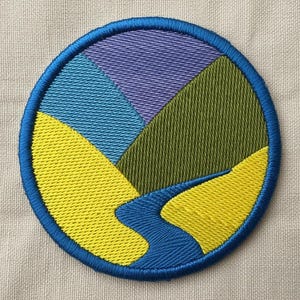 May include: Embroidered patch with a circular design depicting a landscape. The patch has a blue border, with sections of yellow, green, blue, and purple. A blue river meanders through the yellow landscape.