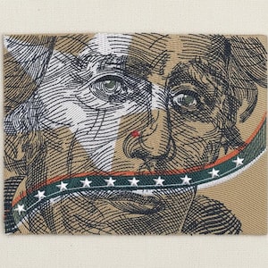 Puede incluir: Un parche de tela beige con una ilustración detallada del rostro de un hombre, con una cinta verde, blanca y roja sobre la boca. El diseño incluye líneas negras y blancas y un punto rojo en la nariz.