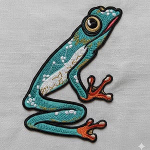 May include: Embroidered patch featuring a turquoise frog with a detailed design. The frog has a white belly, gold accents, and red and orange feet. The patch is outlined in black and appears to be on a light gray fabric.