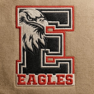 Puede incluir: Parche bordado con la cabeza de un águila estilizada dentro de un diseño de la letra "E". El parche tiene una combinación de colores negro, blanco y rojo, con la palabra "EAGLES" bordada en rojo en la parte inferior. El fondo es de tela color beige.